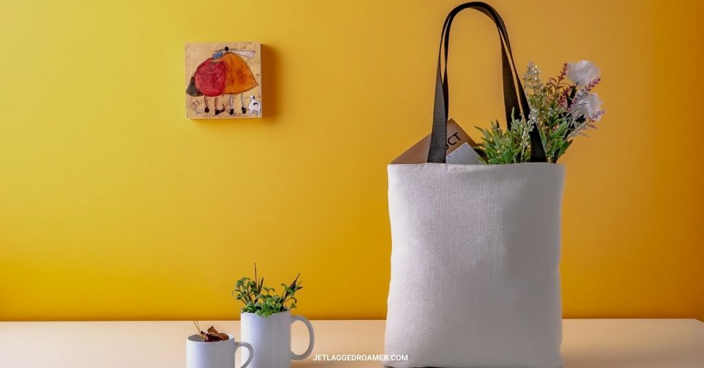 Recyclable bag filled with flowers and other groceries on a table in front of a bright yellow wall. 