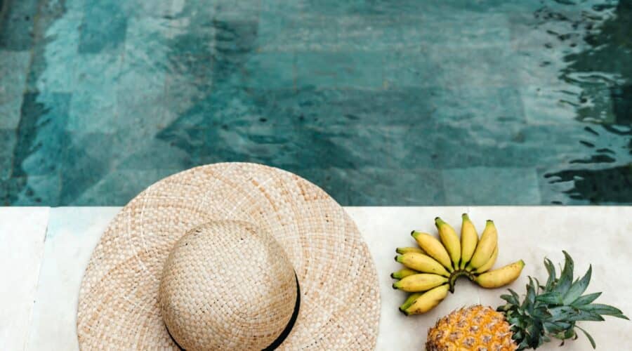 Summer captions picture of a sun hat, bananas, and a pineapple next to a clear water pool.