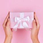 Gift for travel lovers photo os a woman holding a gift with pink wrapping paper and a white bow.