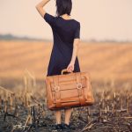 Captions for travel photo of a woman holding a suitcase in an open field alone.